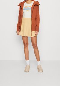 Felpa marrone con zip sopra una t-shirt crema con una grafica di un cuore colorato, abbinata a una gonna chiara plissettata e scarpe chunky beige.