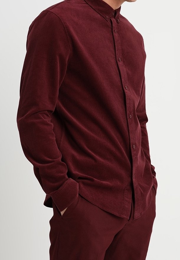 Homme portant une chemise en velours côtelé rouge foncé boutonnée à manches longues avec un pantalon assorti rouge foncé, mains dans les poches sur un fond uni.