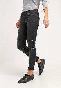 Jeans negros ajustados con detalles de cremallera en las rodillas, diseño de cinco bolsillos y bajos remangados. Combinados con zapatillas negras y camiseta de manga larga gris.