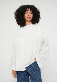 Camisa oversized branca com riscas, de mangas compridas, usada por cima de uma t-shirt branca lisa, combinada com jeans azuis com padrão de diamantes.