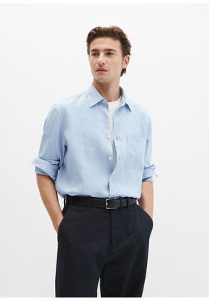 Mann trägt ein hellblaues Hemd mit aufgerollten Ärmeln, ein weißes Unterhemd, einen schwarzen Gürtel und schwarze Hose, die Hände in den Taschen, blickt zur Seite.