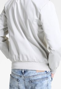 Osoba w jasnoszarej kurtce i niebieskich dżinsach, stojąca z rękami w kieszeniach, zwrócona plecami do kamery.