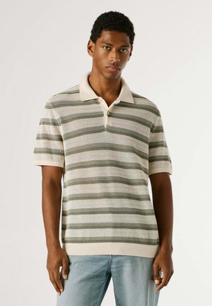 Giovane uomo che indossa una polo a maniche corte a righe verdi e bianche in maglia e jeans azzurri, in piedi davanti a uno sfondo uniforme.