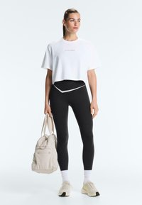 Femme debout tenant un sac de sport beige, portant un T-shirt court blanc avec texte, des leggings noirs avec un détail en forme de V, des chaussettes blanches et des baskets beiges.