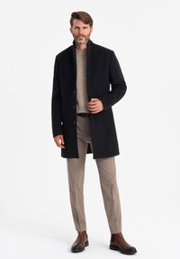Abrigo negro con cuello alto, diseño de un solo botón y tres botones. Combinado con un suéter beige y pantalones de color claro.