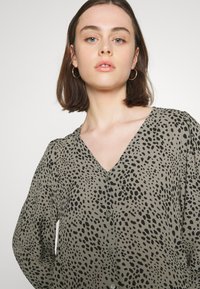 Langermet bluse i myk, lett stoff med en beige bunn og svarte prikker. V-hals design og knappedetaljer foran.