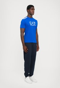 Blue cotton t-shirt with white printed logo, paired with navy joggers. Model wears white sneakers. Simple, fitted design with short sleeves.