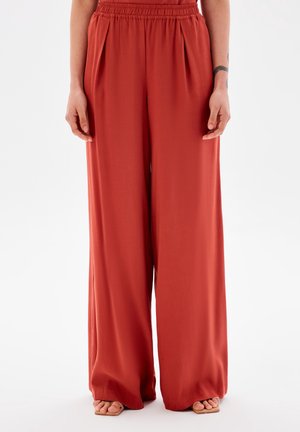 Femme portant un pantalon taille haute à jambes larges de couleur rouille avec une ceinture élastique et des poches, debout pieds nus sur fond blanc.