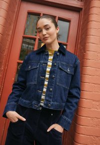 Mørkeblå denimjakke med kort design, features to forlommer, sølvknapper og en spidse krave. Modellen bærer en stribet top.