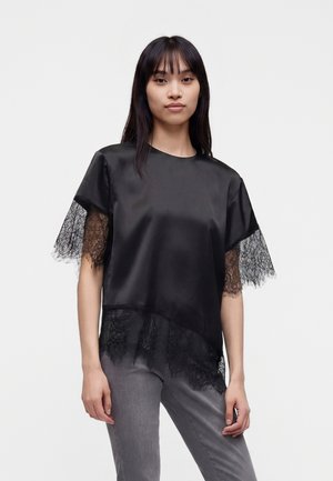 Jeune femme portant un chemisier en satin noir avec manches et ourlet en dentelle, associé à un pantalon gris, debout devant un fond uni.