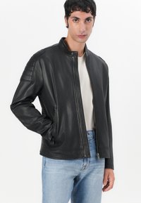 Chaqueta de cuero negra con cuello alzado, cierre frontal y bolsillos laterales. Presenta hombros acolchados y una textura suave. Se lleva sobre una camisa beige.