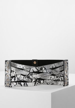 Large ceinture noire et blanche à motif peau de serpent avec des lanières croisées et des clous dorés à chaque extrémité, fermée par un bouton-pression central.