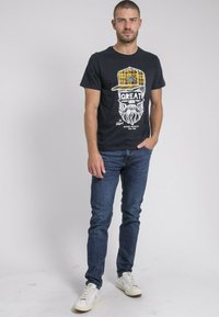 T-shirt noir à manches courtes avec un graphique blanc représentant une barbe et un bonnet à carreaux jaunes. Porté avec un jean bleu foncé et des baskets blanches.