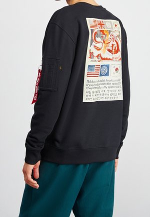 Person iført en sort sweatshirt med et stort farverigt vintage-inspireret patch med flag, en drage og flersproget tekst på ryggen.