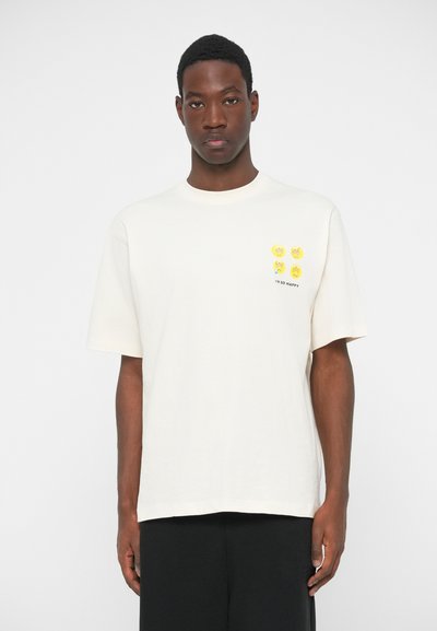 Cream-colored cotton t-shirt with relaxed fit. Features yellow emoji graphics on the chest and black text reading "I'M SO HAPPY."