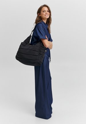 Zwarte puffer tas met doorgestikte textuur en ritssluitingdetails, gedragen over de schouder. Gecombineerd met een blauw stoffen outfit met wijde pijpen.
