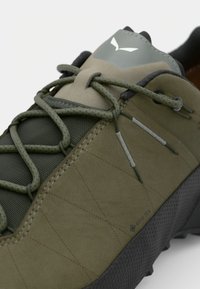 Zöld túracipő texturált felülettel, steppelt dizájnnal és megerősített gumitalppal. Gore-Tex logóval és rugalmas fűzőrendszerrel rendelkezik.