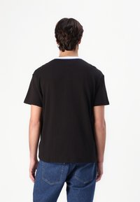 Mies, jolla on lyhyet tummat hiukset, mustassa T-paidassa ja sinisissä farkuissa, näkyy selkäpuolelta yksinkertaista valkoista taustaa vasten.