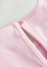 Vaaleanpunainen puuvillapusero, jossa on pyöreä pääntie, takana sydänkuvioinen napitus ja näkyvät sauman yksityiskohdat.