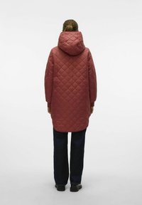 Veste longue matelassée rouge avec une capuche, présentant un motif en losanges et des poignets élastiques, assortie à un pantalon droit bleu marine et des chaussures noires.