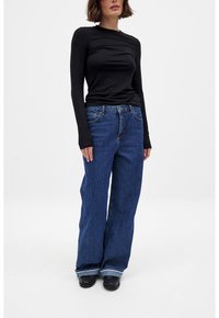 Camiseta negra de manga larga combinada con vaqueros anchos de color azul, con una textura de denim clásica y un dobladillo ligeramente desgastado. Lleva zapatos negros.