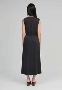 Femme aux longs cheveux noirs portant une robe mi-longue noire sans manches et des sandales à talons bruns, se tenant de dos sur un fond gris.