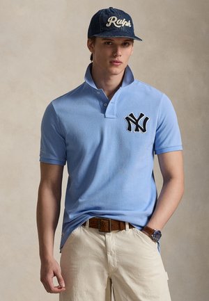 Jasnoniebieska koszulka polo z podniesionym kołnierzykiem i czarnym haftowanym logo NY, w połączeniu z beżowymi szortami i jeansową czapką z napisem "Ralph" w stylu pisma odręcznego.