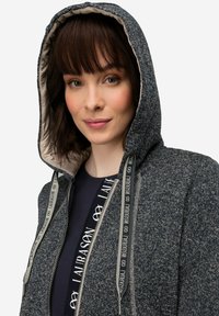 Grå huvtröja med ett texturerat tyg och ett kontrasterande ljus inre foder, med dekorativa snören längs dragkedjan och en framficka.