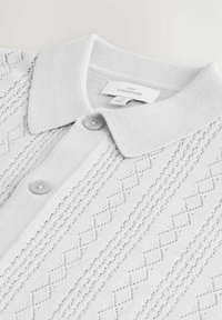 Camiseta polo blanca con un patrón de diamantes texturizado, que cuenta con un cuello de canalé, dos botones y una etiqueta de tela tejida en el interior.