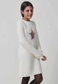 Giovane ragazza che indossa un vestito maglione bianco con una stella di paillettes multicolore, in piedi davanti a uno sfondo grigio chiaro uniforme.