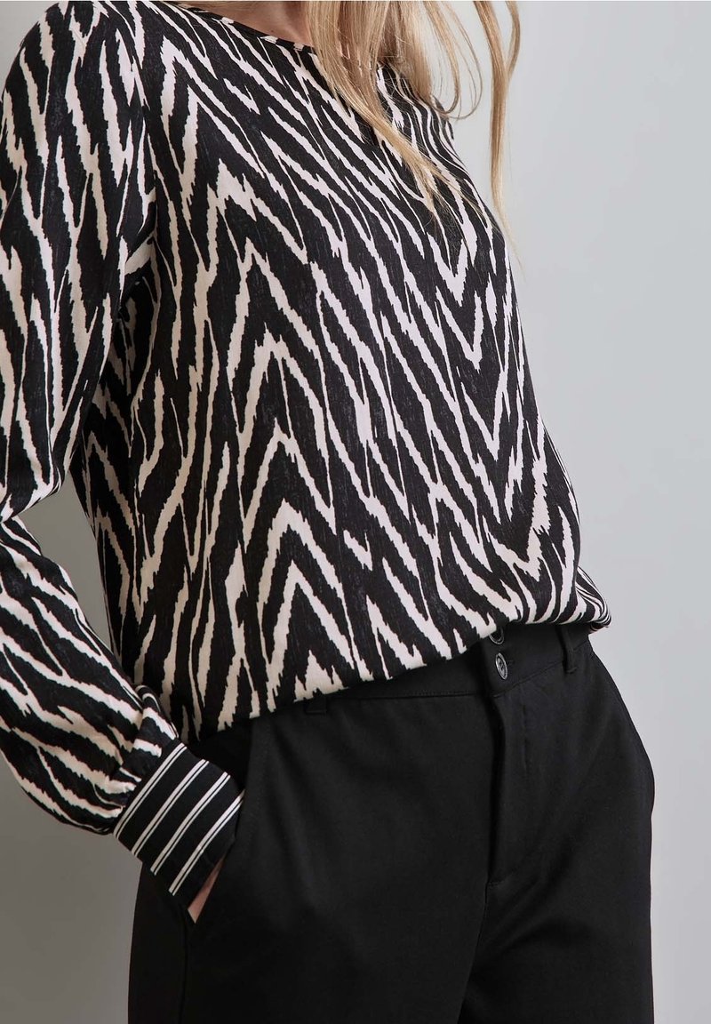 Blouse à motifs noirs et blancs avec des rayures en zigzag, manches longues avec poignets rayés, associée à un pantalon noir.