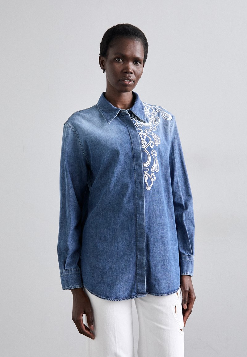 Camicia di jeans a maniche lunghe, color blu sbiadito, con collo, caratterizzata da ricami floreali bianchi sul lato sinistro, indossata con pantaloni bianchi.