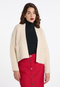 Cremefarvet fluffy cardigan med vid åbning forneden, to forlommer og ribbet kant, parret med en sort turtleneck og rød nederdel.