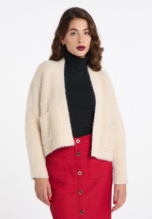 Crème-kleurige fuzzy cardigan met een brede open voorkant, twee voorzakken en ribgebreide afwerking, gecombineerd met een zwarte coltrui en een rode rok.