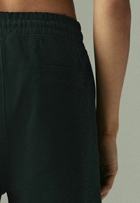 Pantalones cortos negros vistos desde atrás, con una tela texturizada, cinturilla elástica y un pequeño bolsillo de parche en el lado derecho.