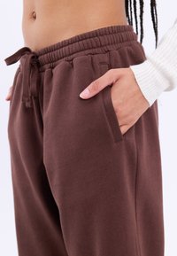 Bruine joggingbroek met elastische tailleband en trekkoord. Beschikt over zijzakken en een zachte textuur, afgebeeld met een hand in een van de zakken.