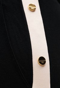 Μαύρο πλεκτό ύφασμα με μια κεντρική λευκή λωρίδα που φέρει ένα χρυσό κουμπί. Το κουμπί έχει μαύρο πάνω μέρος με μια χρυσή γραμμή και ανάγλυφη διακόσμηση.