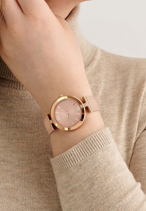 Reloj de oro rosa con una esfera redonda y cara rosa, correa de cuero con acentos decorativos, usado en una mano contra un suéter beige.