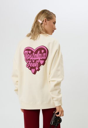 Cremefarbener Sweater mit einem großen, pinken Herz-Graphic und dem Text "Nostalgia Feels", oversized Ärmeln und entspanntem Schnitt.