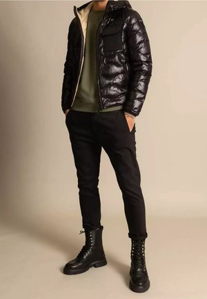 Homme portant une doudoune noire brillante, un sweat-shirt vert olive, un pantalon noir et des bottes de combat noires à lacets, debout les mains dans les poches.