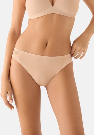 Mujer vistiendo un sujetador beige sin costuras y ropa interior tipo bikini a juego contra un fondo liso y claro.