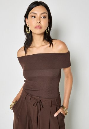 Kobieta ubrana w brązową bluzkę odkrywającą ramiona i pasujące spodnie, z złotymi kolczykami-kołami i nałożonymi na siebie bransoletkami, stojąca na tle jednolitego tła.