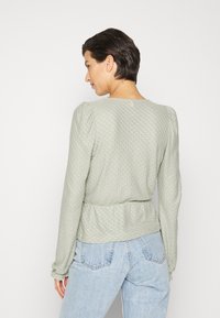 Top de manga longa, verde claro, com um padrão texturizado, cintura franzida e ombros bufantes, combinado com jeans de cintura alta, azul claro.