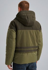 Puffjacka i mörkgrön och olivton, med huva, quiltat mönster och horisontella mörka ränder över ryggen.