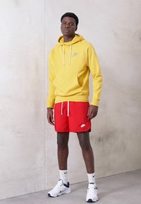 Rumena kapuca s sweatshirtom z vrvicami, rdeče kratke hlače z belim logotipom in bele superge s črnimi dodatki. Manekenka stoji ob nevtralni steni.