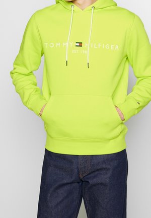 Osoba, která má na sobě limetkově zelenou mikinu Tommy Hilfiger s přední kapsou a bílými tkaničkami, v kombinaci s tmavě modrými džínami.