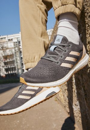 Jambes vêtues d'un pantalon beige et de chaussettes blanches portant des chaussures de course Adidas noires et beiges sur un fond urbain extérieur.