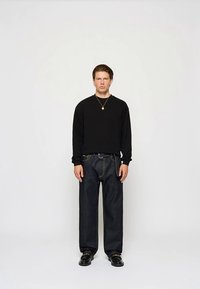 Μαύρο πουκάμισο μακρυμάνικο σε συνδυασμό με σκούρο μπλε, φαρδιά τζιν παντελόνια. Φοράει ένα χρυσό μενταγιόν και μαύρα παπούτσια. Απλός σχεδιασμός.