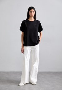Black short-sleeve t-shirt with a round neck and small gold logo, paired with wide-legged white pants and white sneakers, set against a gray background.