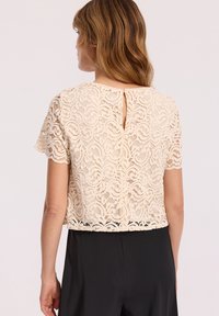 Koronkowy top w kolorze kremowym z kwiecistymi wzorami, krótkimi rękawami i detalem w kształcie dziurki z tyłu. Połączony z czarnymi spodniami o wysokim stanie.